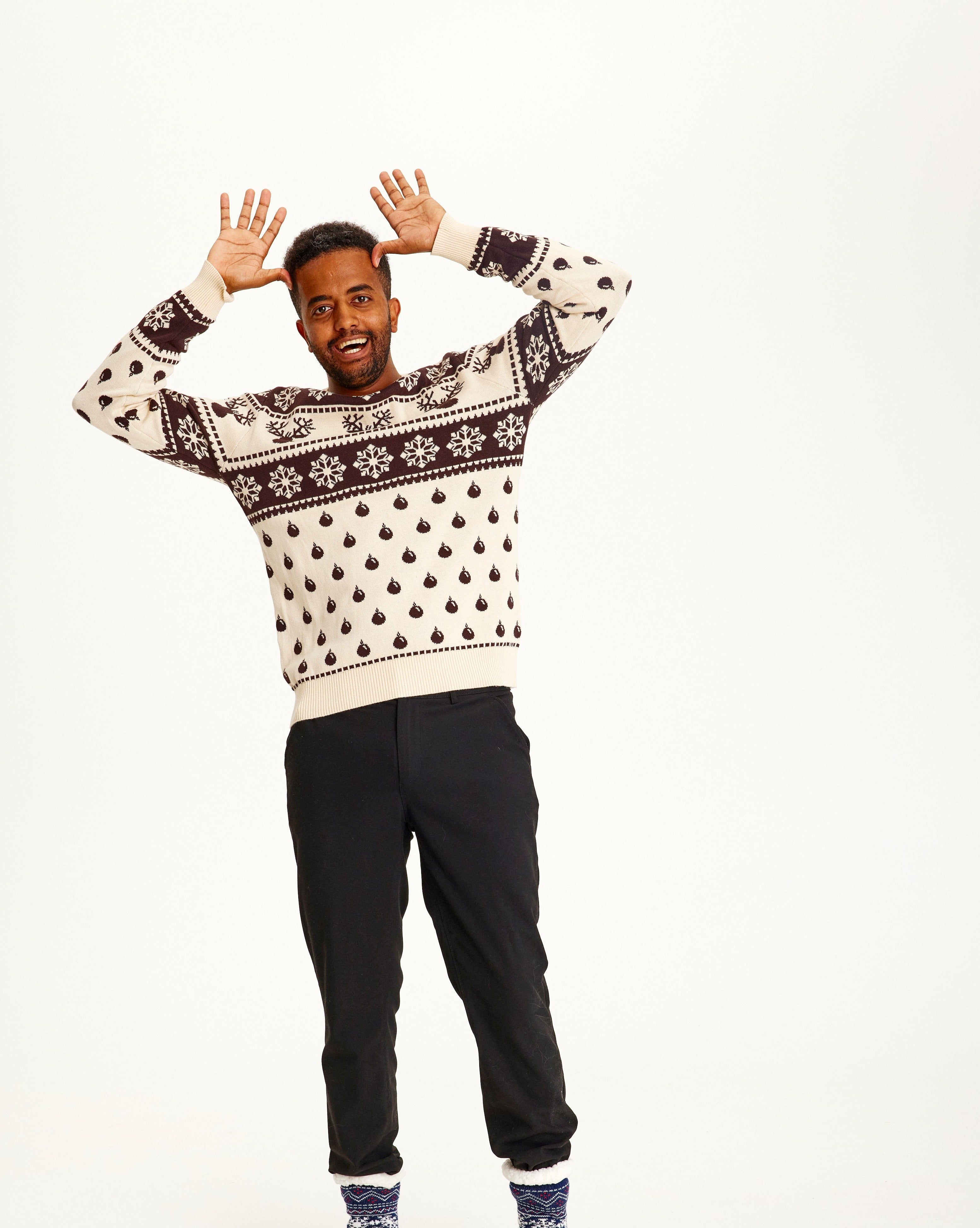 Un homme qui pose, portant un pull de Noël beige et marron avec des flocons de neige et des motifs.